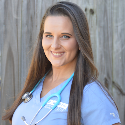 A dark haired veterinary assistant with seal blue scrubs smiles brightly at the camera.